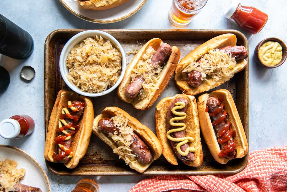 A top down shot of beer brats on buns with mustard and ketchup on a baking sheet with a small bowl of sauerkraut.