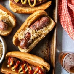 A top down shot of beer brats with sauerkraut on buns with mustard and ketchup on a baking sheet.