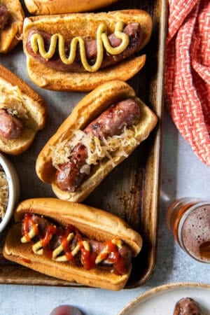 A top down shot of beer brats with sauerkraut on buns with mustard and ketchup on a baking sheet.