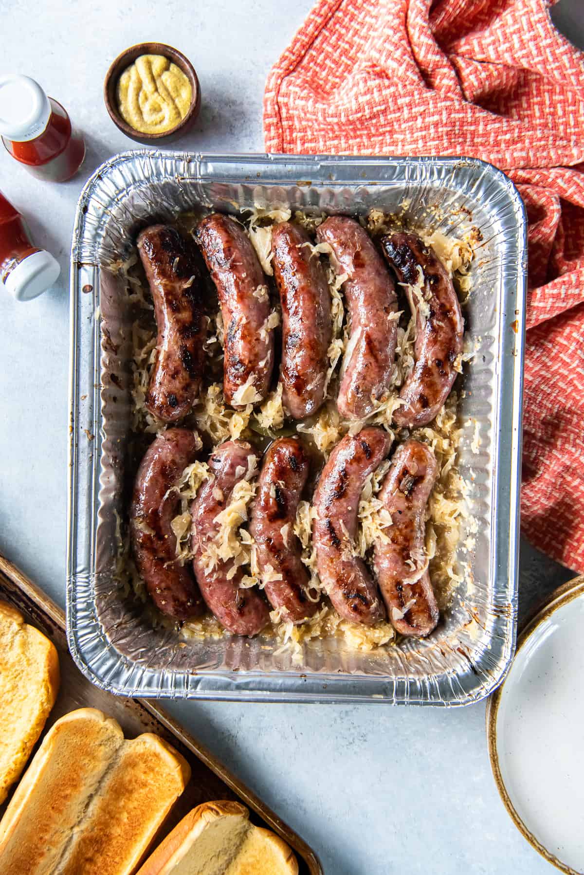 Beer brats with sauerkraut in a foil pan next to toasted hot dog buns and a small bowl of mustard.