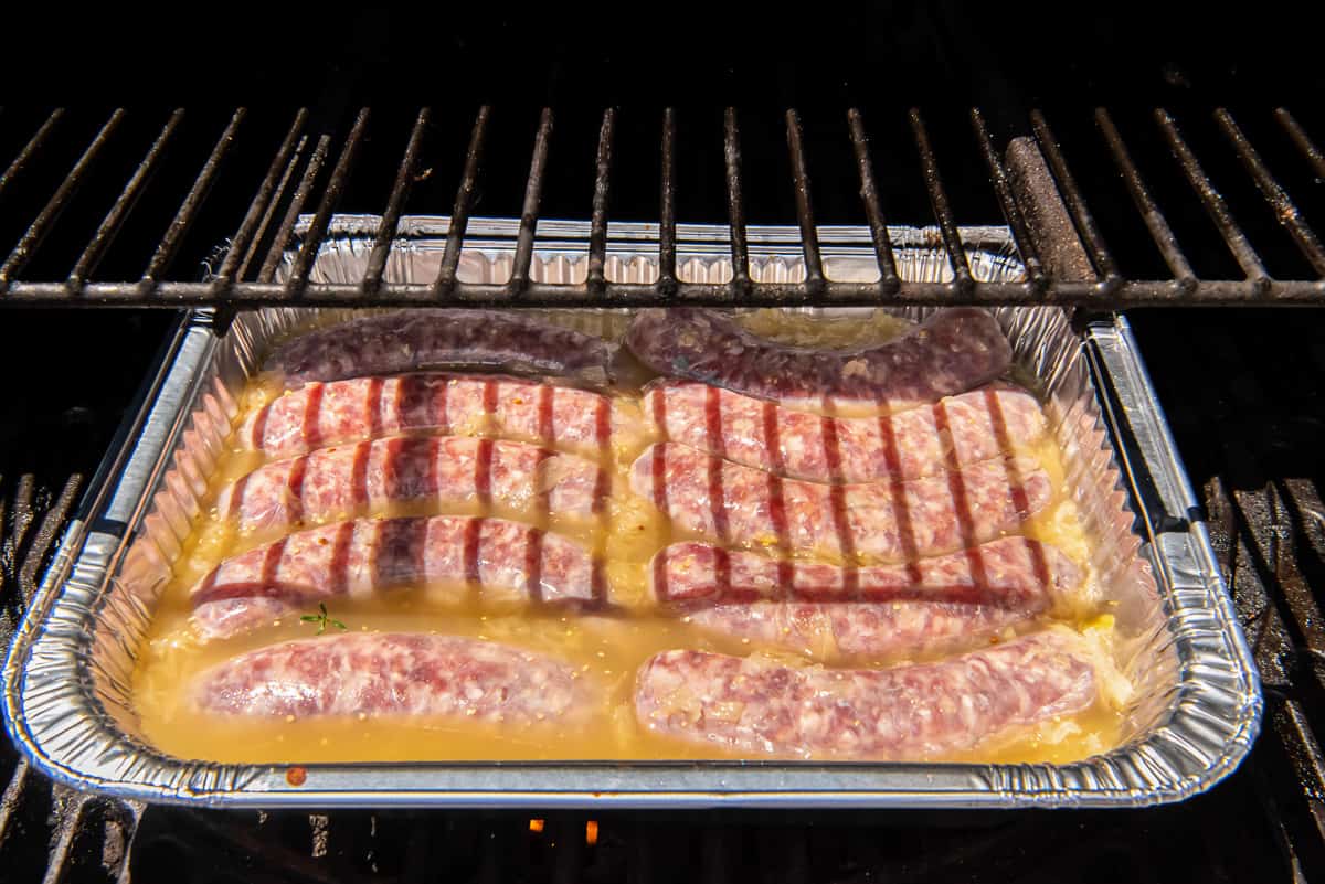 Bratwurst and sauerkraut submerged in beer in a foil pan cooking on a grill.