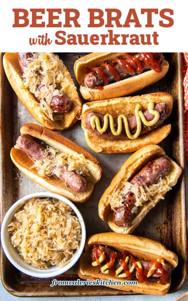 A top down shot of beer brats on buns with mustard and ketchup on a baking sheet with a small bowl of sauerkraut with text.