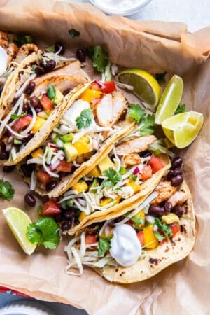 A top down shot of chicken tacos with sour cream, cheese and mango salsa on parchment paper.