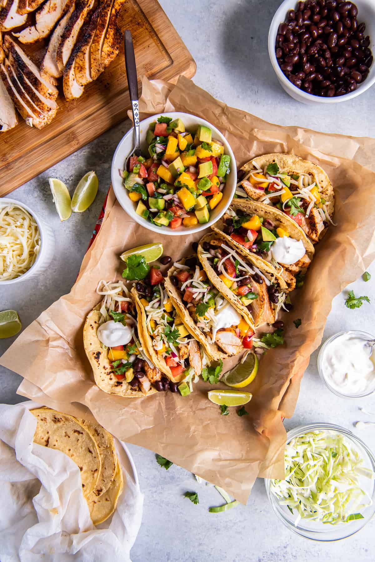A top down shot of chicken tacos topped with mango salsa on parchment paper surrounded by bowls of ingredients.