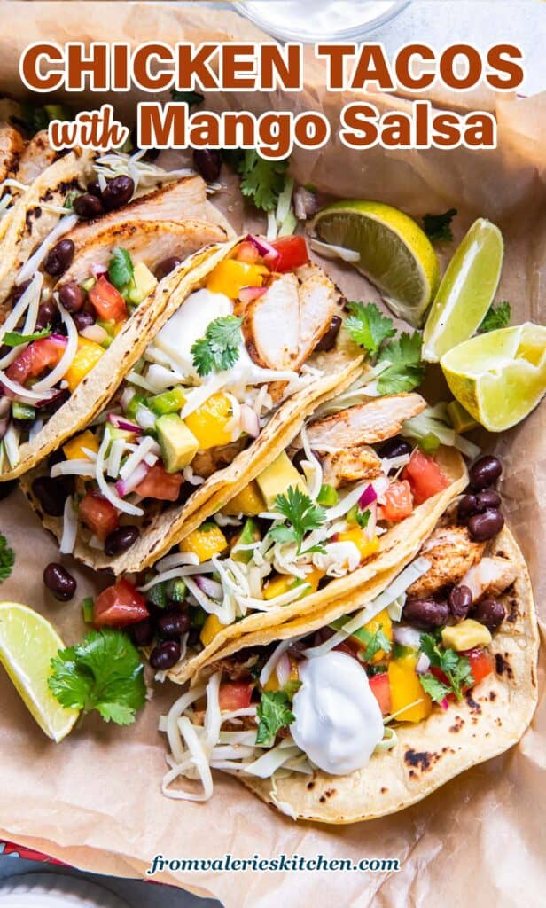 A top down shot of chicken tacos with sour cream, cheese and mango salsa on parchment paper with text.