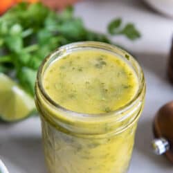 A mason jar filled with cilantro lime vinaigrette with cilantro and vegetables behind it.