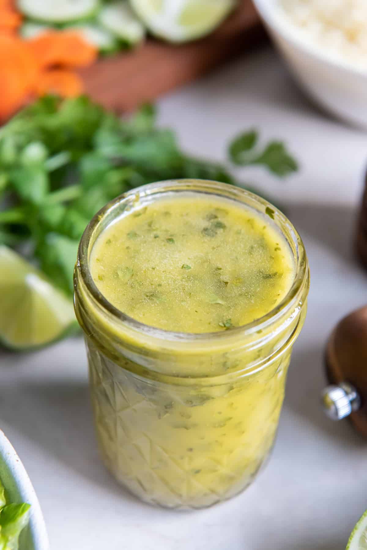 A mason jar filled with cilantro lime vinaigrette with cilantro and vegetables behind it.