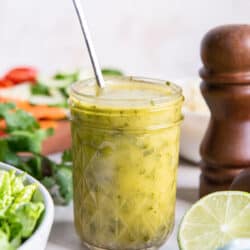 A spoon resting in a mason jar filled with cilantro lime vinaigrette on a kitchen counter with salad ingredients.