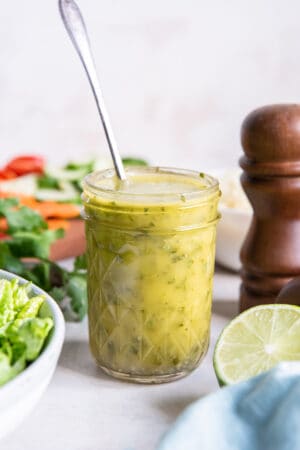 A spoon resting in a mason jar filled with cilantro lime vinaigrette on a kitchen counter with salad ingredients.