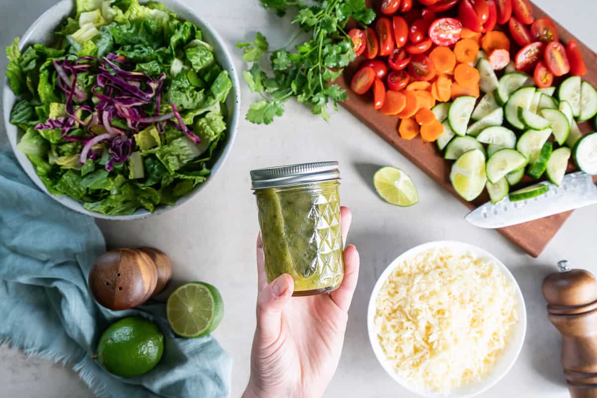 A top down shot of a hand holding a mason jar filled with cilantro lime vinaigrette with salad and vegetables underneath it.