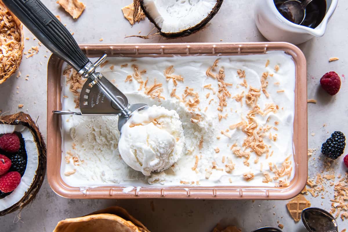 A top down shot of an ice cream scoop resting in a metal loaf pan filled with coconut ice cream.