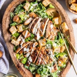 A top down shot of two salad spoons resting in a wood bowl filled with grilled chicken Caesar salad.
