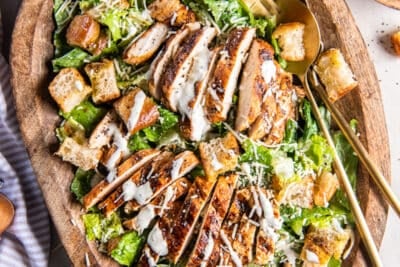 A top down shot of two salad spoons resting in a wood bowl filled with grilled chicken Caesar salad.