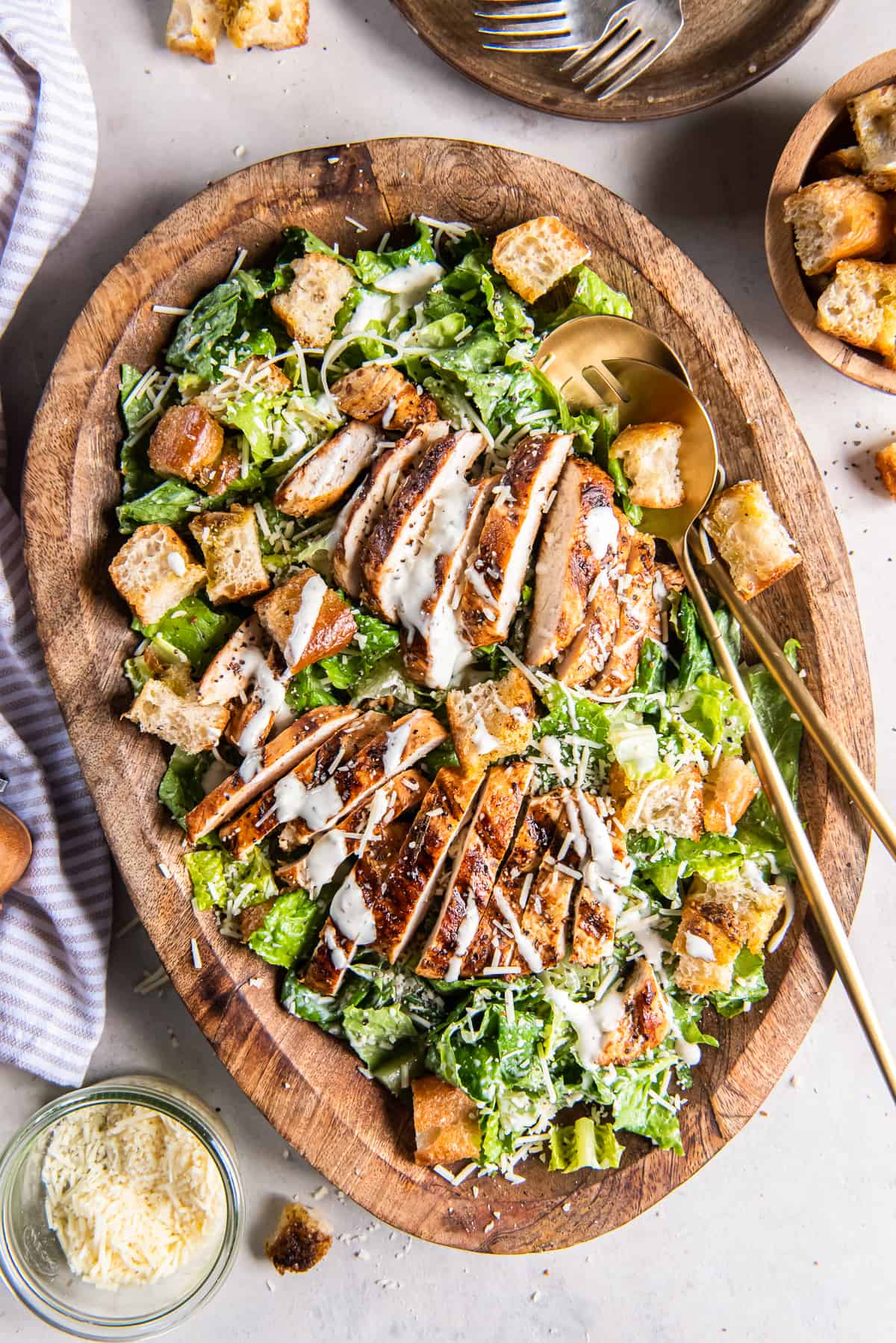 A top down shot of two salad spoons resting in a wood bowl filled with grilled chicken Caesar salad.