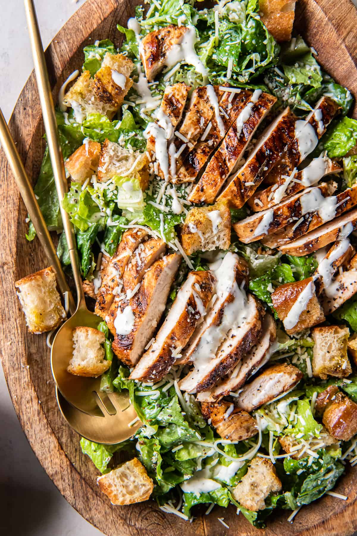A top down close up shot of two salad spoons resting in a wood bowl filled with grilled chicken Caesar salad.
