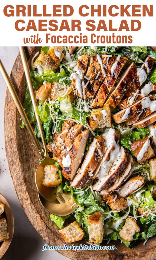 A top down shot of salad spoons resting in a Caesar salad with grilled chicken and focaccia croutons in a wood bowl with text.
