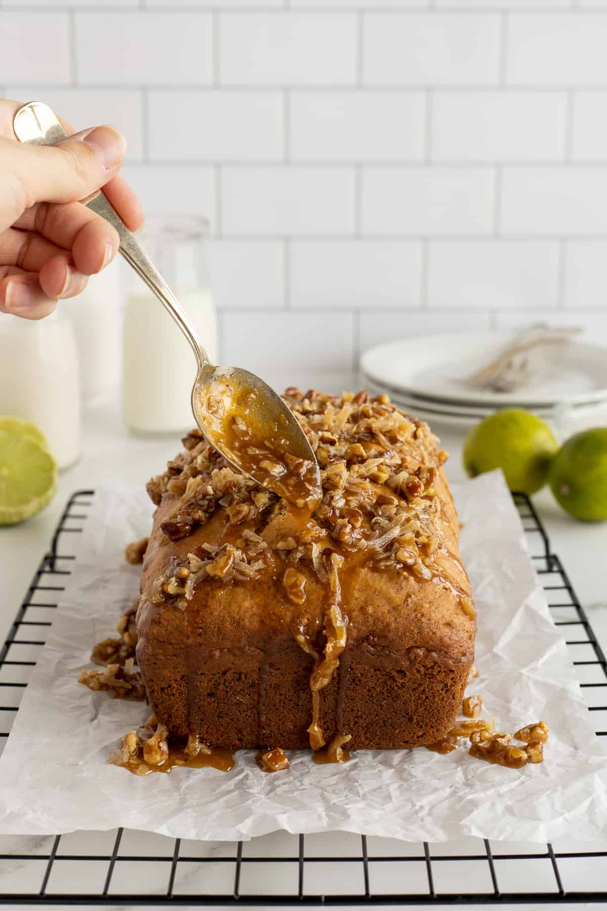 A hand drizzling a coconut pecan mixture from a spoon over a loaf of banana bread.