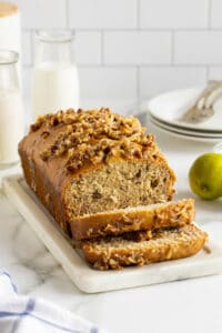 A partially sliced loaf of banana bread topped with a coconut pecan mixture on a rectangular plate.