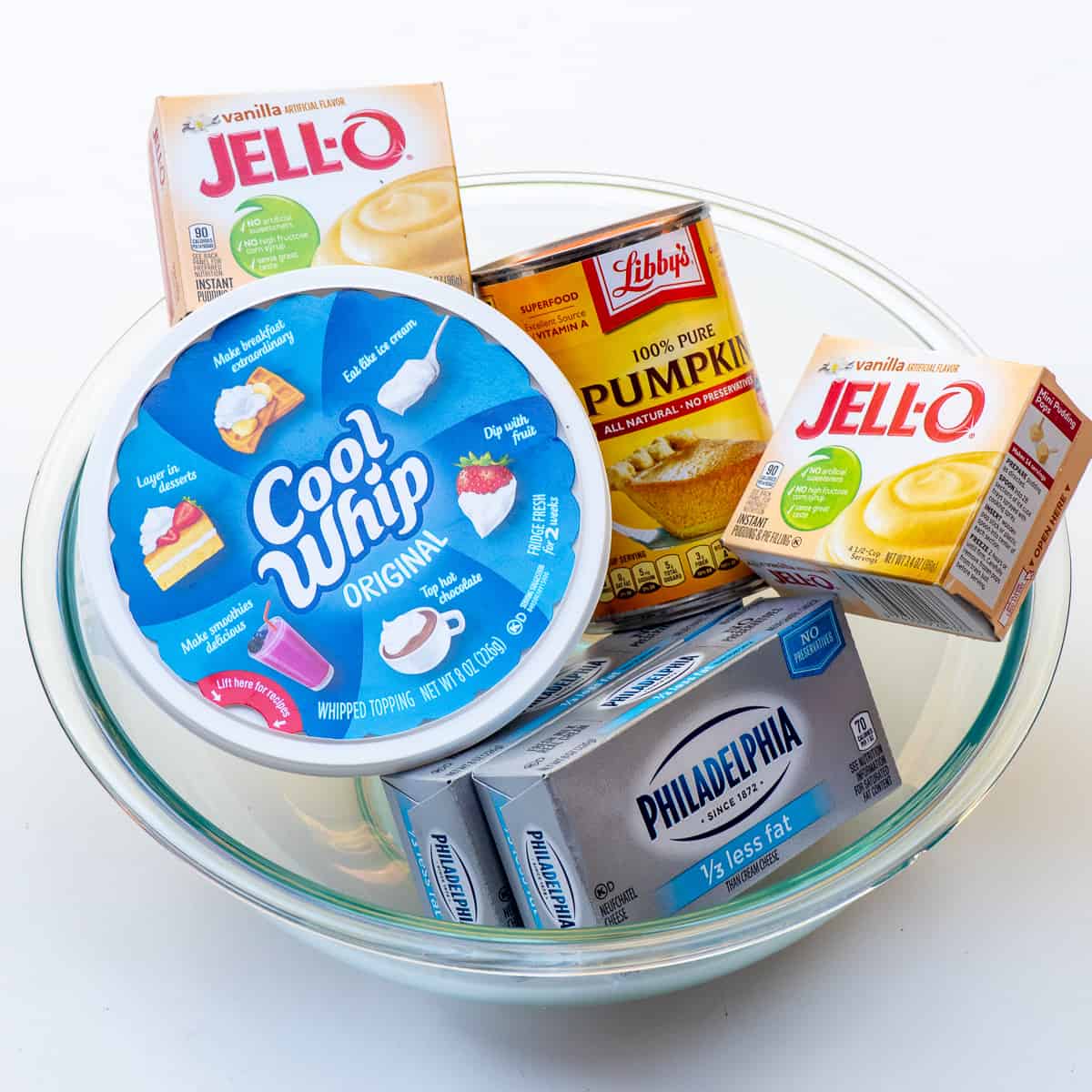 A tub of Cool Whip, can of pumpkin puree, two boxes of Jello pudding and two boxes of cream cheese in a glass bowl.