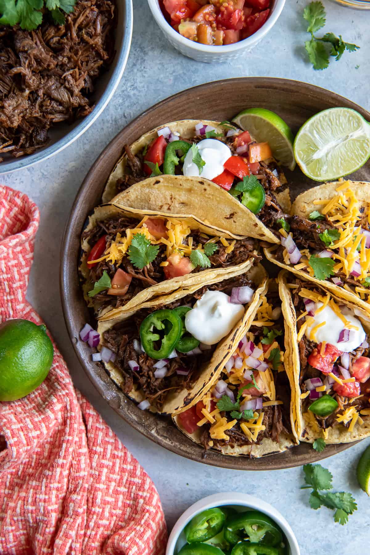 A top down shot of a platter of shredded beef tacos with toppings and lime wedges.