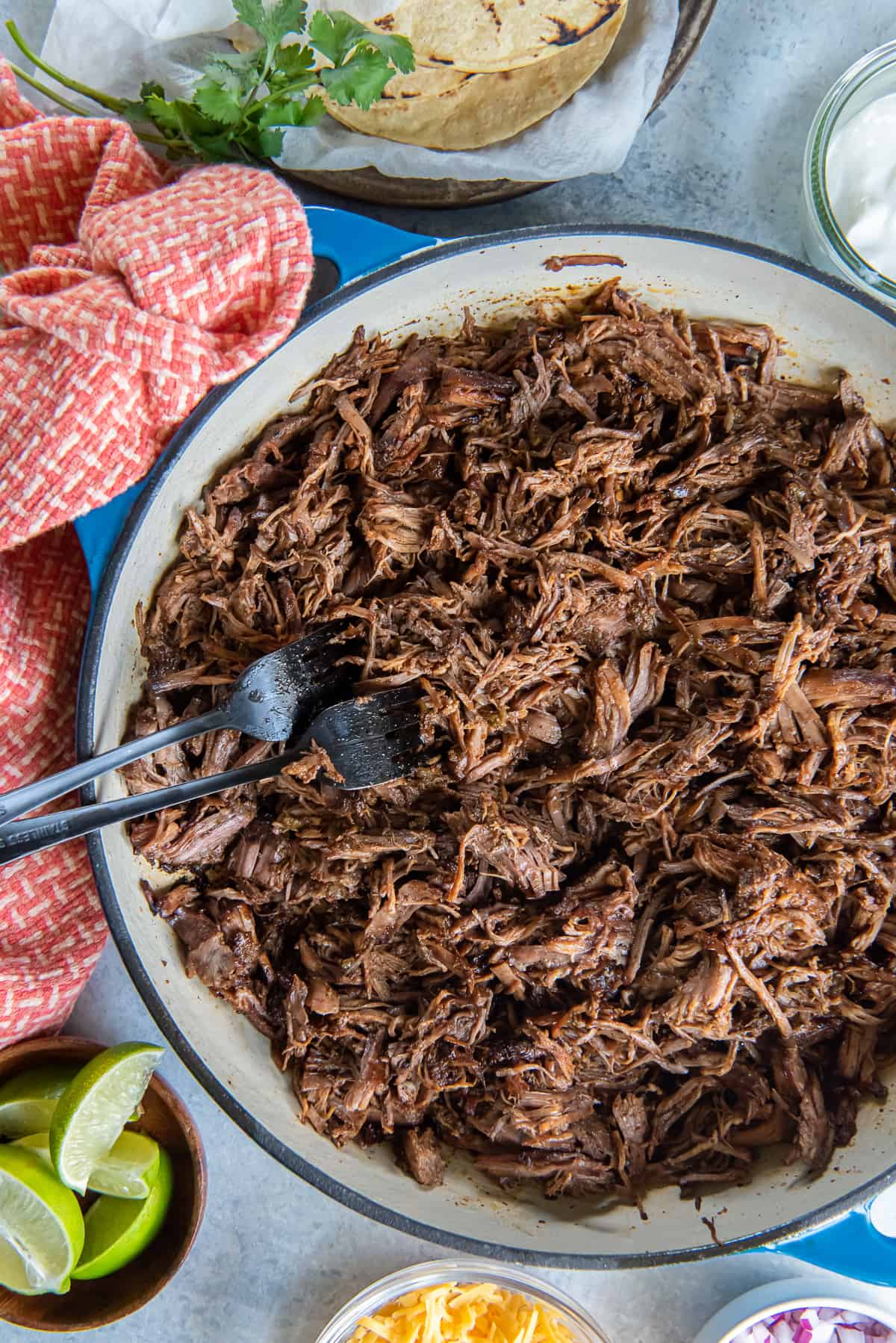 A top down shot of two forks resting in a pan full of Mexican shredded beef surrounded by bowls of taco toppings.