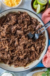 A top down shot of two forks resting in a pan full of Mexican shredded beef surrounded by bowls of taco toppings.