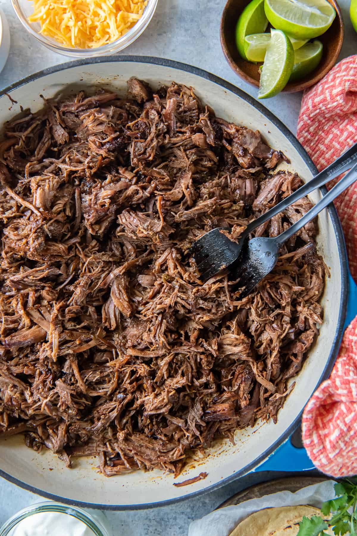 A top down shot of two forks resting in a pan full of Mexican shredded beef surrounded by bowls of taco toppings.