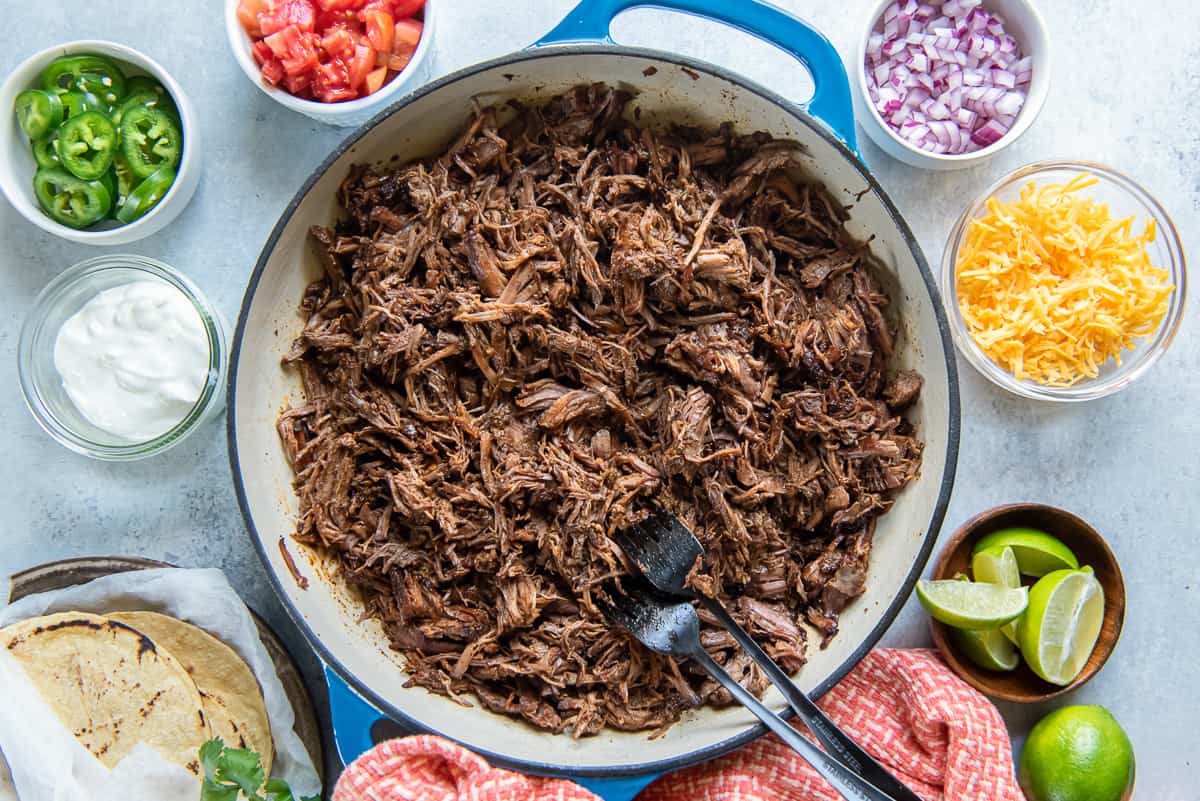 A top down shot of two forks resting in a pan full of Mexican shredded beef surrounded by bowls of taco toppings.