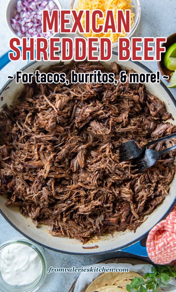 A top down shot of two forks resting in a pan full of Mexican shredded beef surrounded by bowls of taco toppings with text.