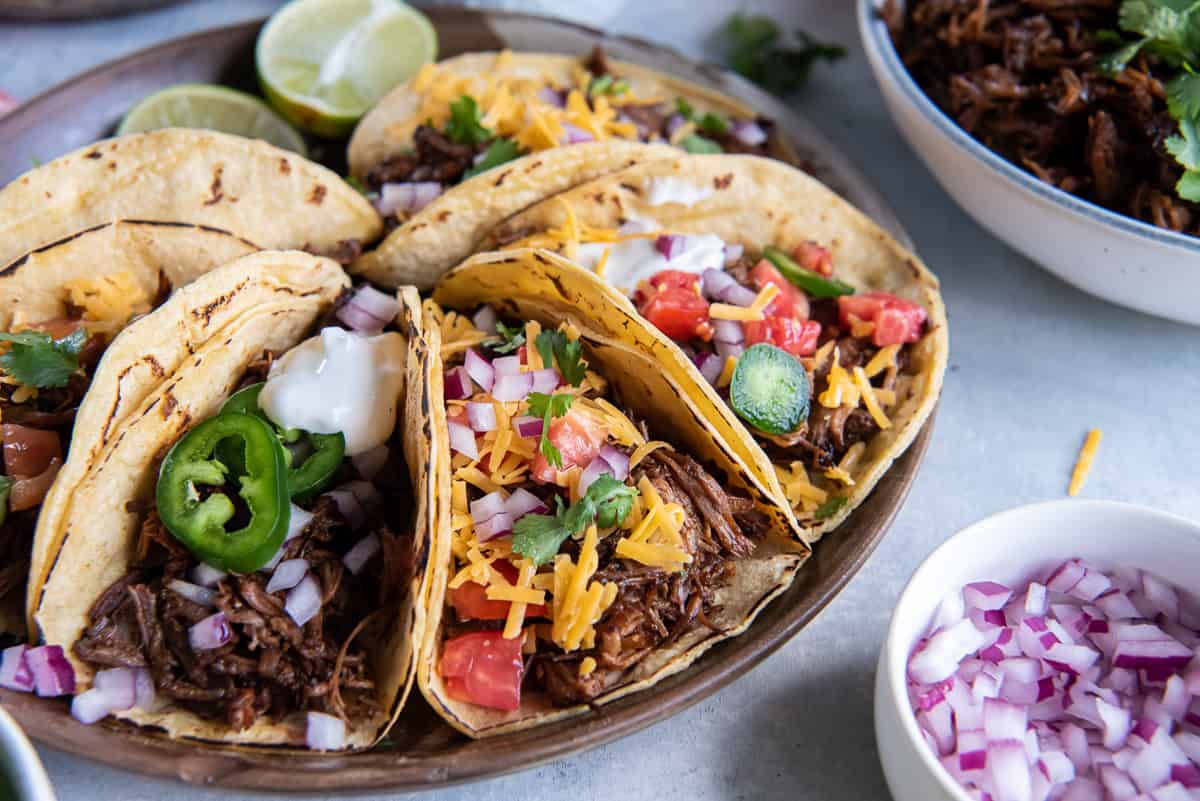 Shredded beef tacos with cheese, tomato, and other toppings on a wood platter.