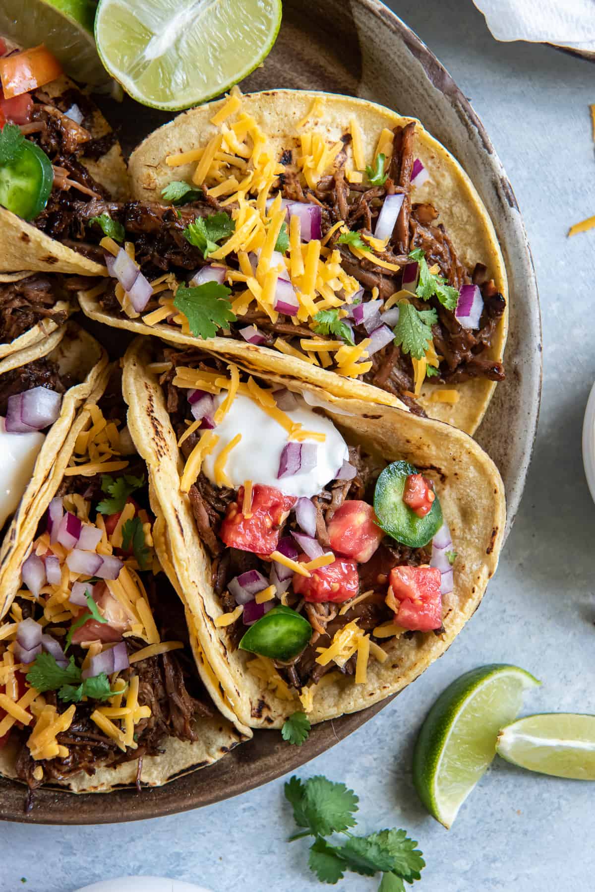 A top down shot of a platter of shredded beef tacos with toppings and lime wedges.