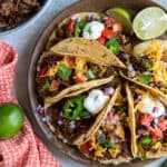 A top down shot of a platter of shredded beef tacos with toppings and lime wedges.