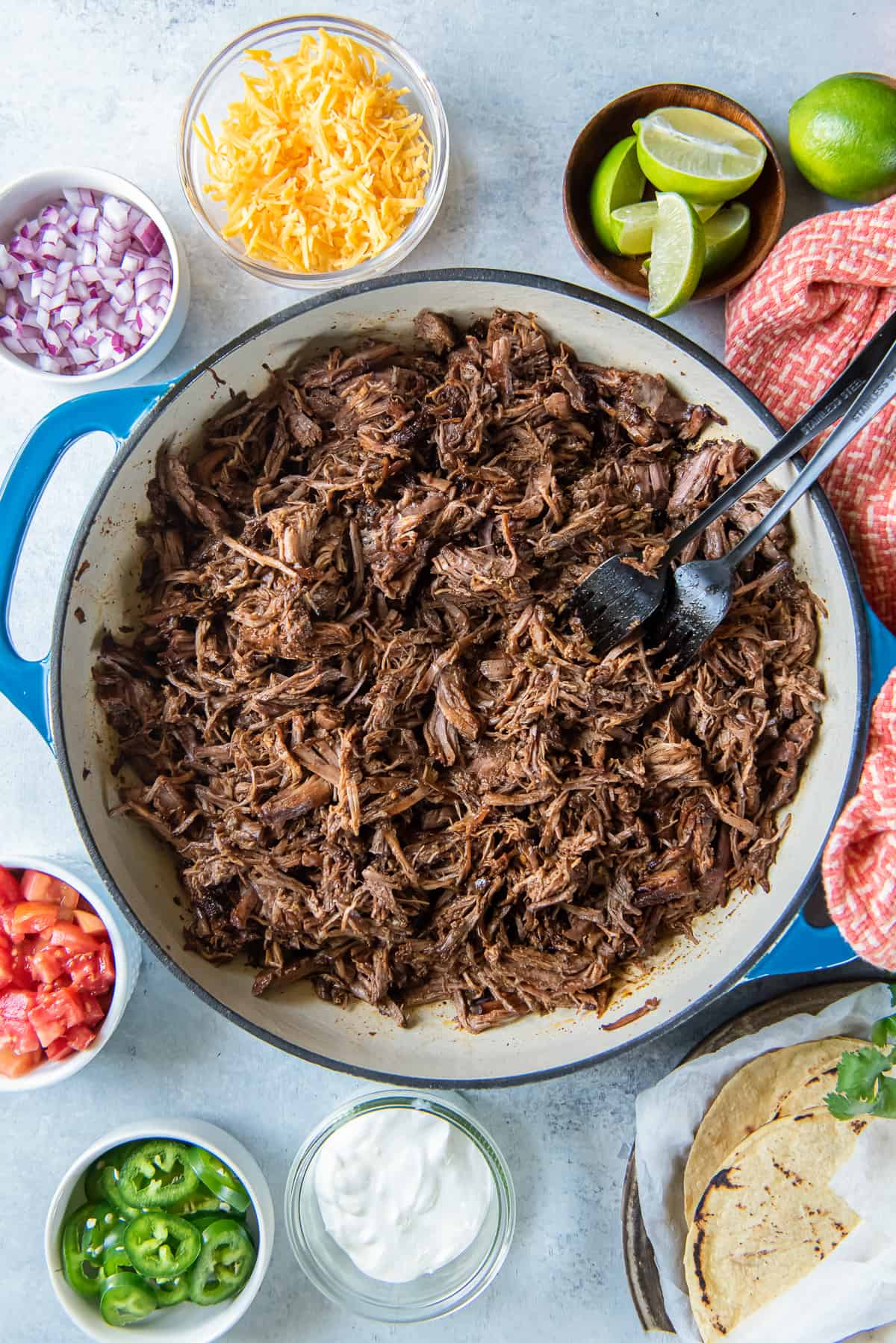 A top down shot of shredded beef in a pan surrounded by taco toppings in bowls and corn tortillas.