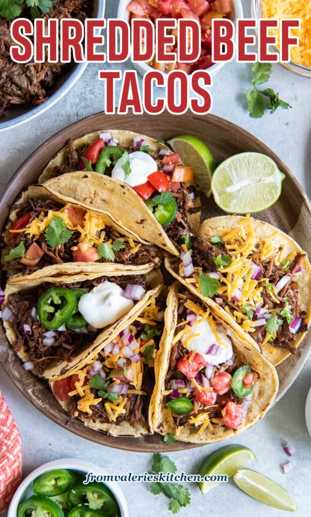 A top down shot of a platter of shredded beef tacos with toppings and lime wedges with text.