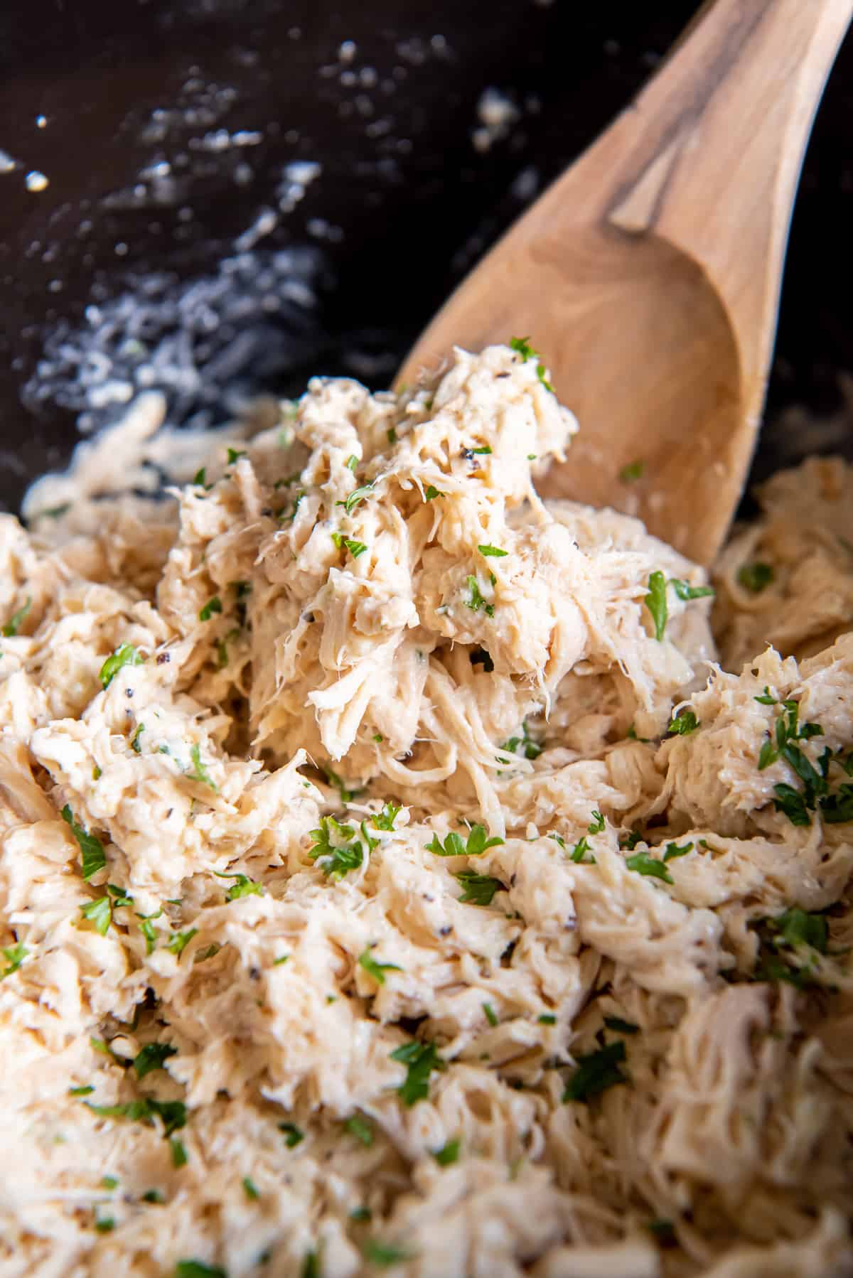 A close up of a wooden spoon scooping shredded Caesar chicken from a slow cooker.