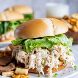 A bun stuffed full with shredded Caesar chicken and romaine lettuce on a white plate with pretzel snack mix.