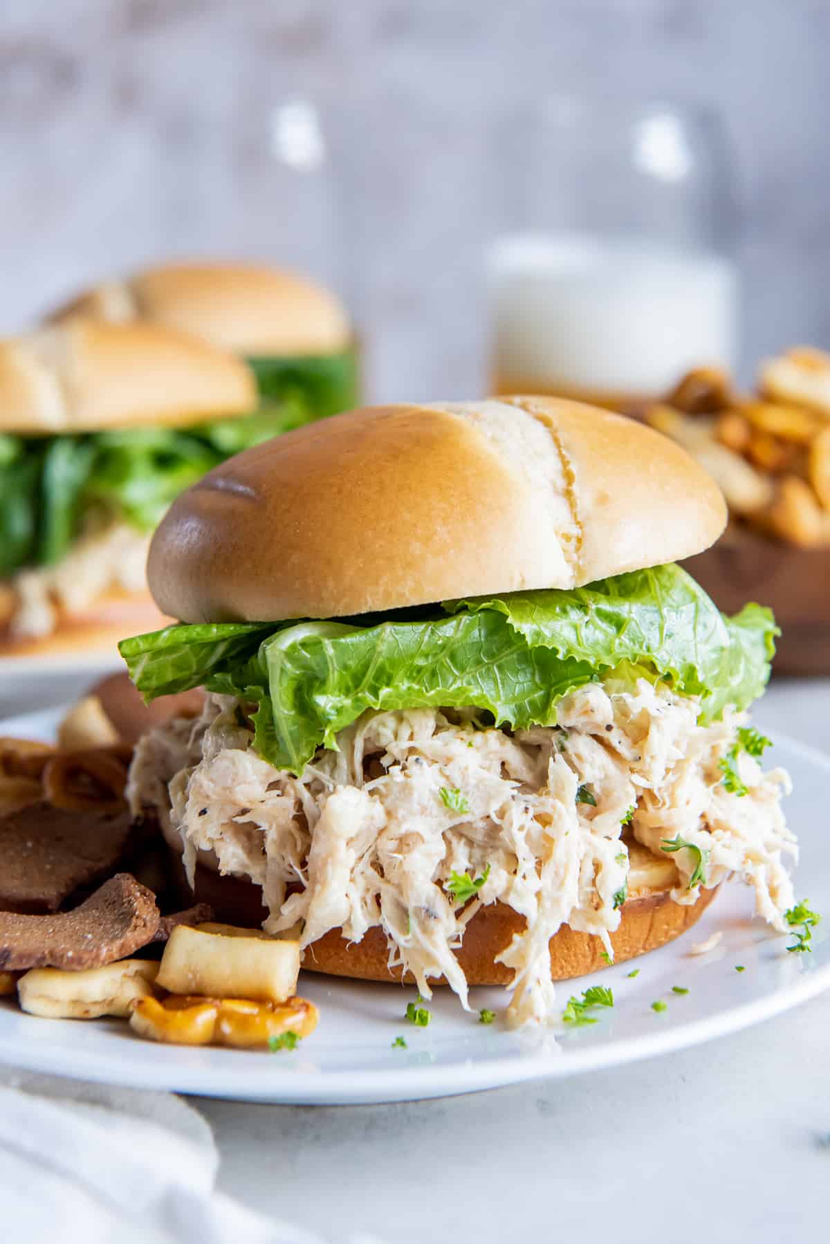 A bun stuffed full with shredded Caesar chicken and romaine lettuce on a white plate with pretzel snack mix.