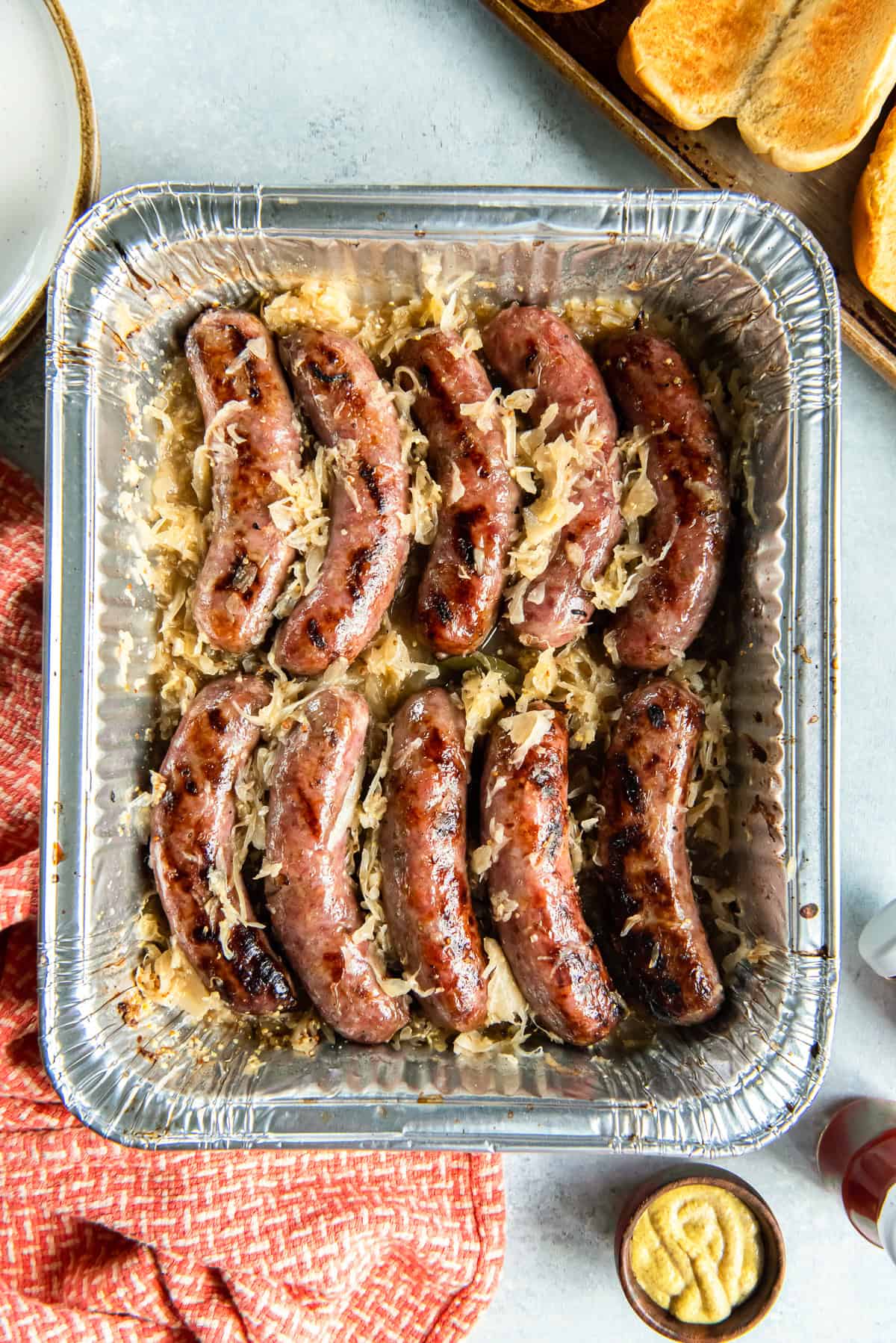 Beer brats with sauerkraut in a foil pan next to toasted hot dog buns and a small bowl of mustard.