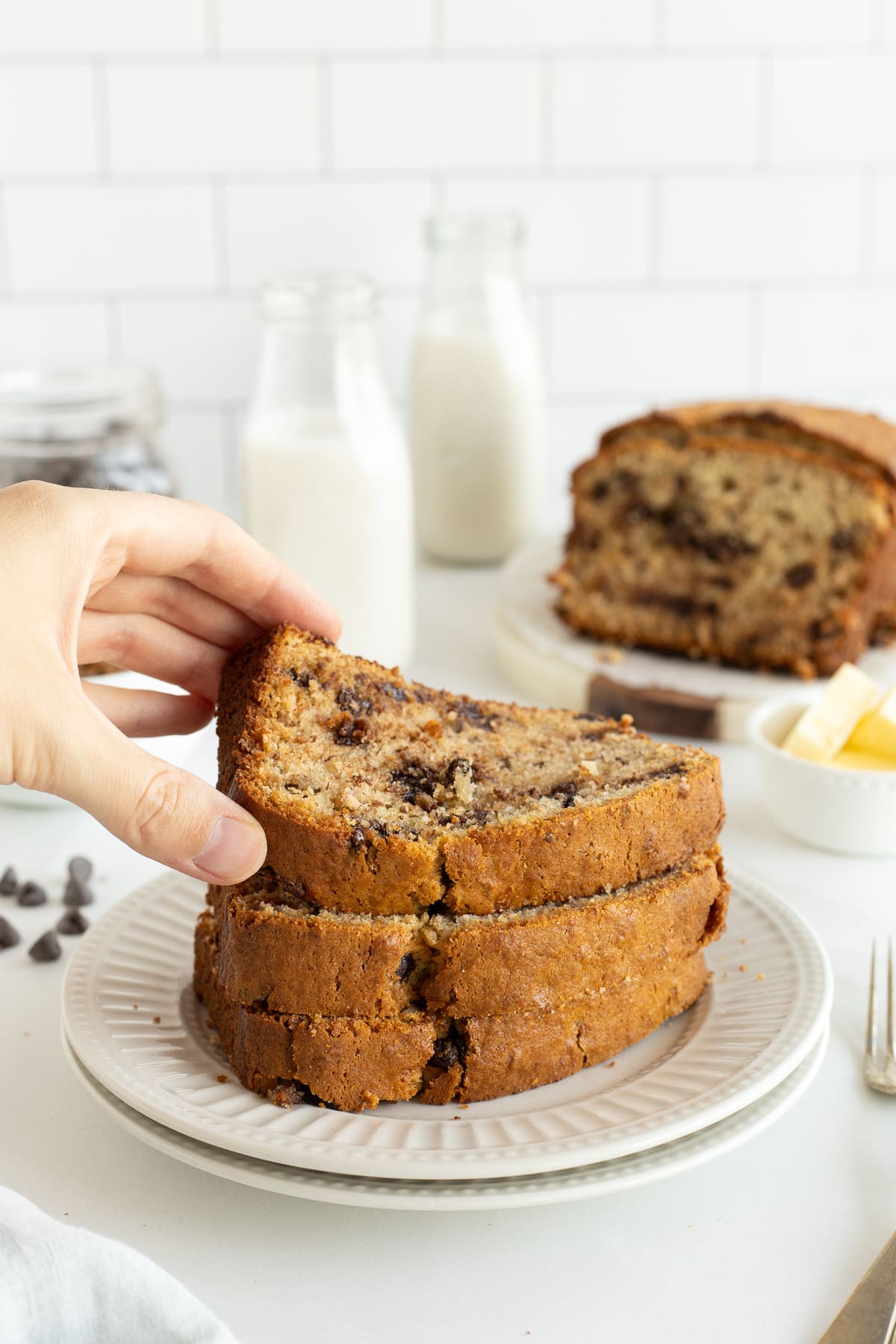 A hand lifting a slice of chocolate chip banana bread off a stack of three slices.