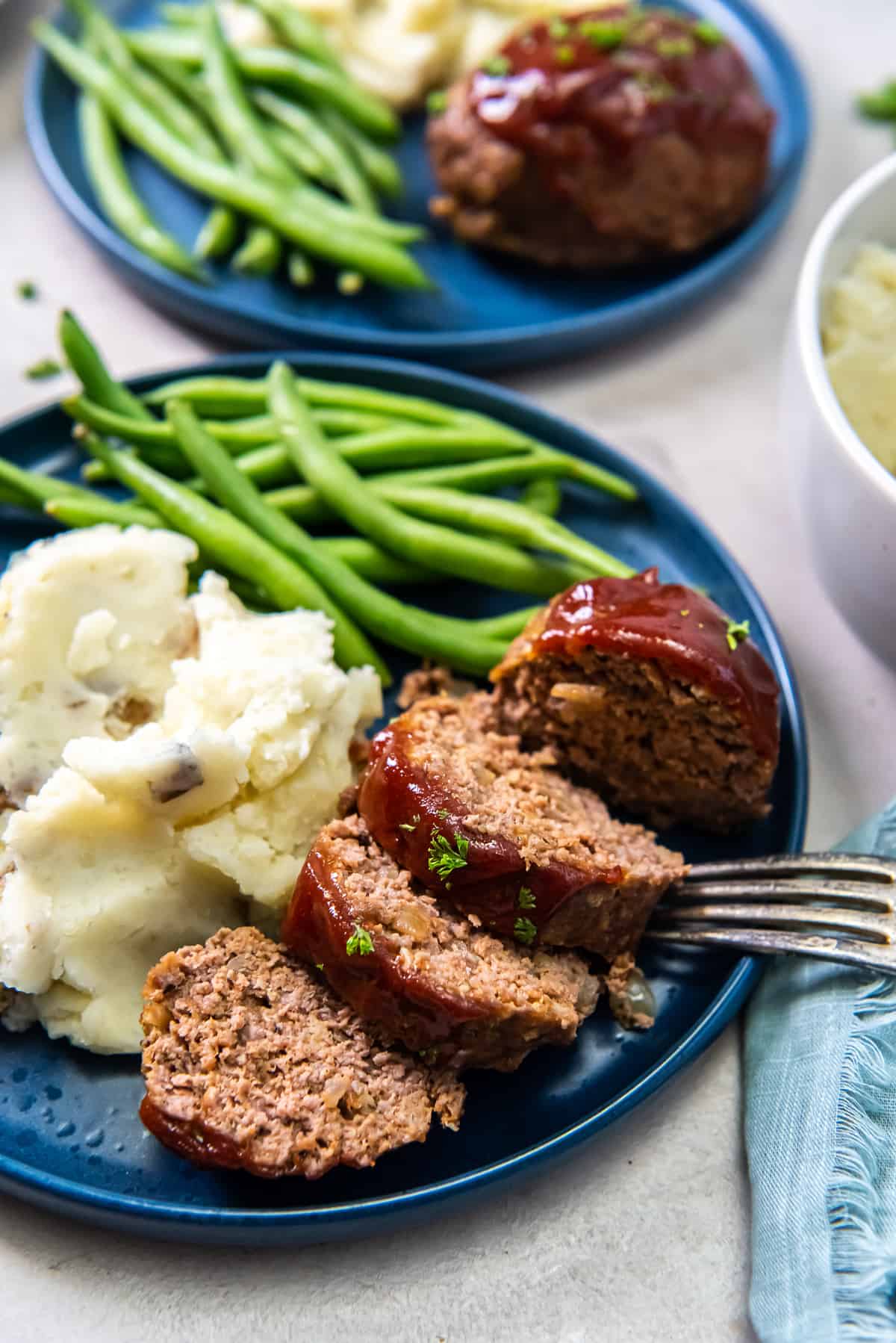Slices of meatloaf on a blue plate with mashed potatoes and green beans.