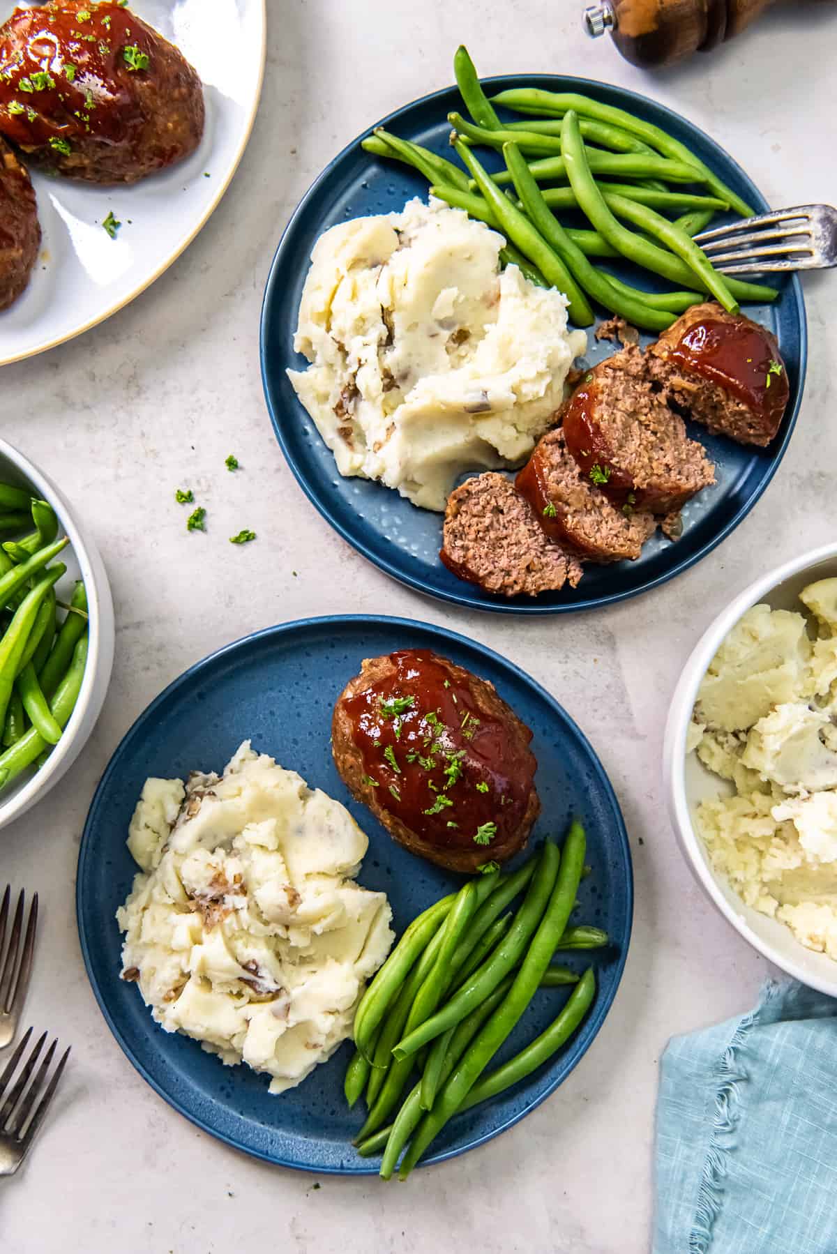 A top down shot of two blue plates with mini meatloaf, mashed potatoes, and green beans.
