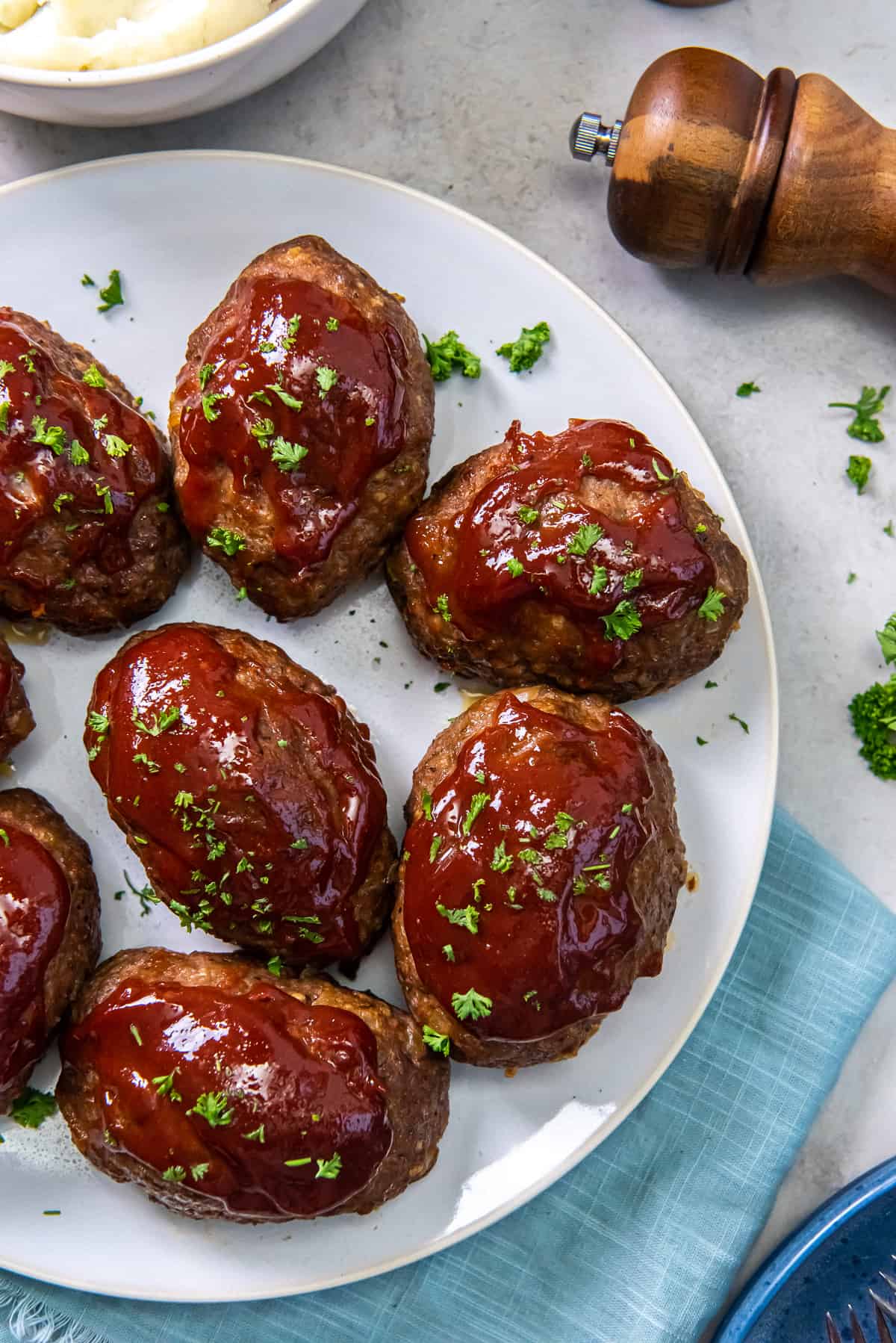 A top down shot of glazed mini meatloaves on a white plate next to a blue cloth and a pepper grinder. 