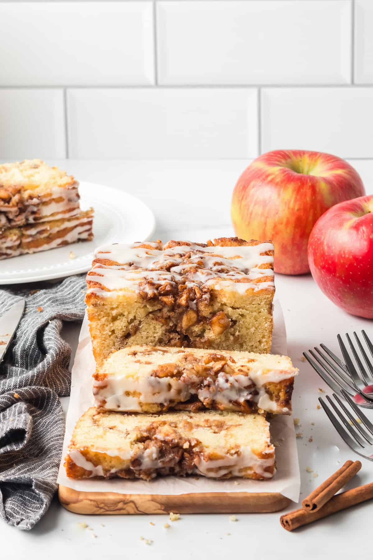 A sliced loaf of quick bread with apples and glaze on a wood board on a kitchen counter with apples in the background.