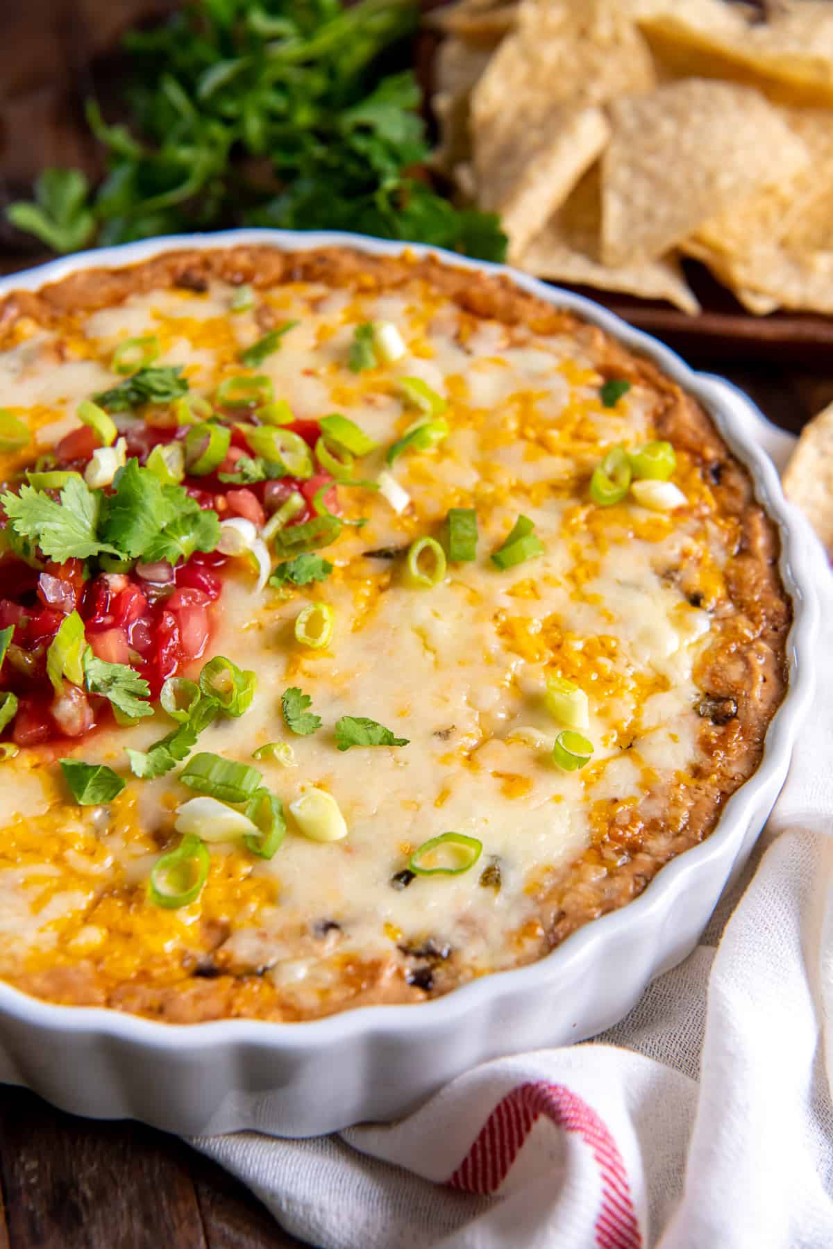 A close up of cheesy bean dip in a white baking dish on a white and red kitchen towel.