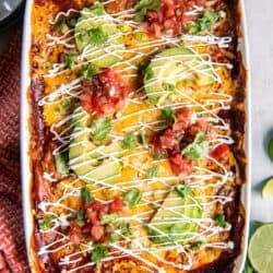 A top down shot of a baking dish filled with chicken enchilada casserole topped with sliced avocado and sour cream on an orange and white kitchen towel.