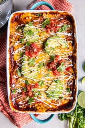 A top down shot of a baking dish filled with chicken enchilada casserole topped with sliced avocado and sour cream on an orange and white kitchen towel.