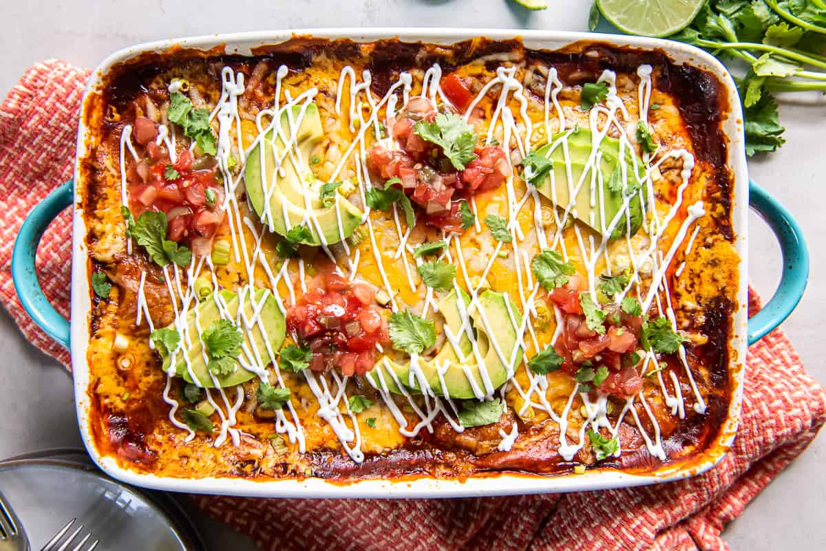 A top down shot of a baking dish filled with chicken enchilada casserole topped with sliced avocado and sour cream on an orange and white kitchen towel.