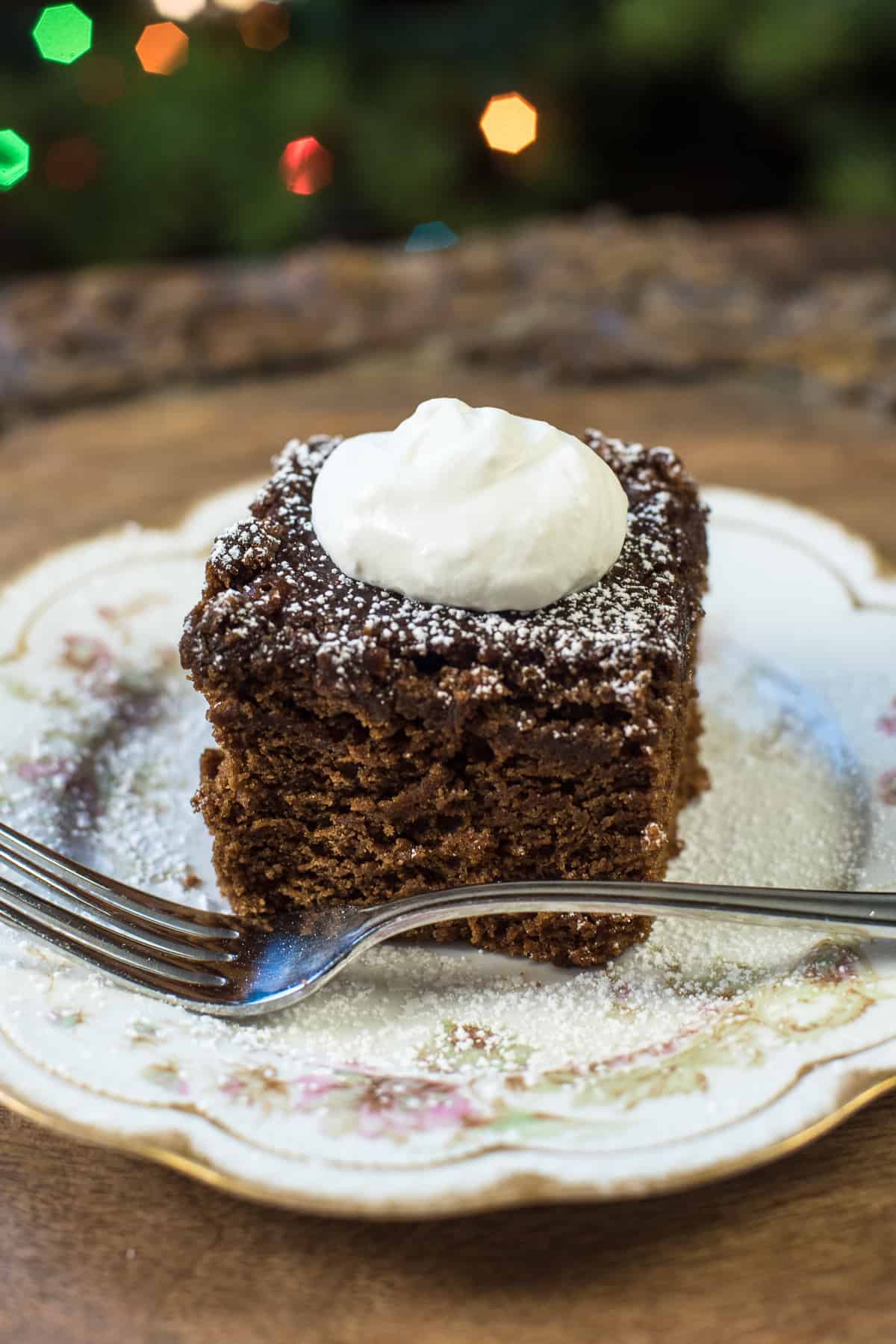 A slice of gingerbread topped with powdered sugar and whipped cream on a pretty china plate with a fork.