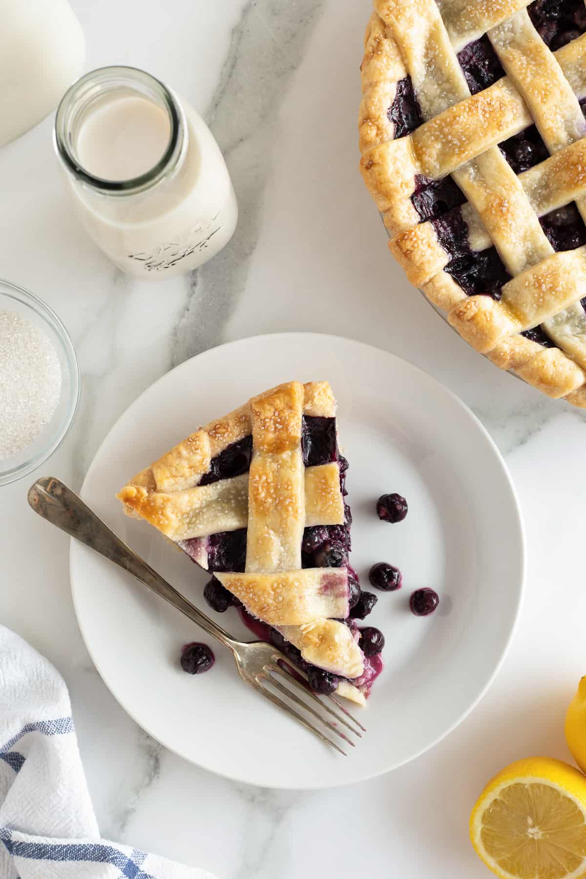 A top down shot of a slice of blueberry pie with lattice crust next to a pie dish.