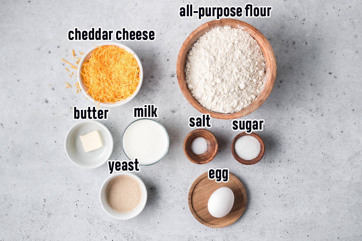 Shredded cheddar cheese, flour, yeast and other ingredients in bowls with text.